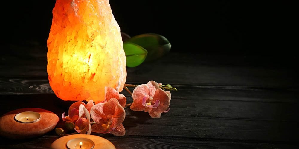Himalayan salt lamp, candles and flowers on table against dark b
