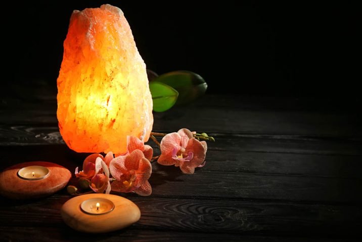 Himalayan salt lamp, candles and flowers on table against dark b